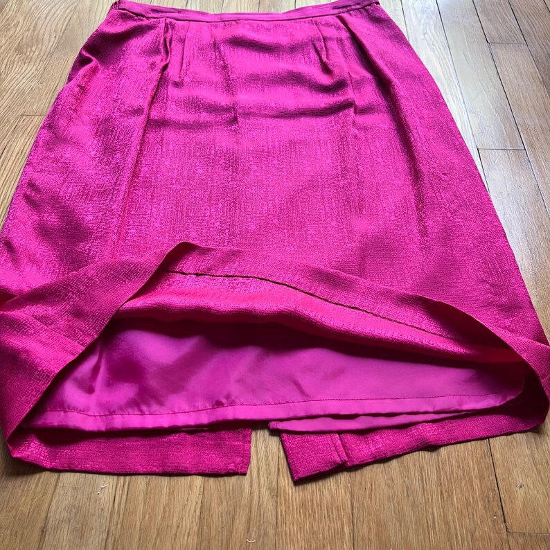 Peut inclure: Une jupe rose vif avec un tissu textur&eacute; et une fente &agrave; l'ourlet. La jupe a une taille ajust&eacute;e et un ourlet au genou. La doublure est rose fonc&eacute;. La jupe est faite d'un mat&eacute;riau brillant.