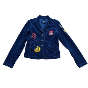 May include: A navy blue corduroy blazer featuring a notched lapel, three front buttons, and two flap pockets. The jacket is decorated with various embroidered patches, including a red number 3, a yellow circular patch, and mountain-themed emblems.