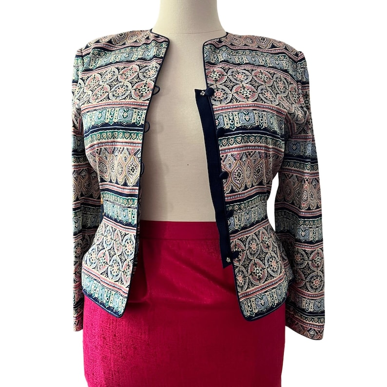 Peut inclure: Veste &agrave; motifs avec bordure et boutons bleu marine. La veste pr&eacute;sente un motif r&eacute;p&eacute;titif dans les tons rose, bleu et beige. Elle est associ&eacute;e &agrave; une jupe rouge vif. La veste est pr&eacute;sent&eacute;e sur un mannequin.