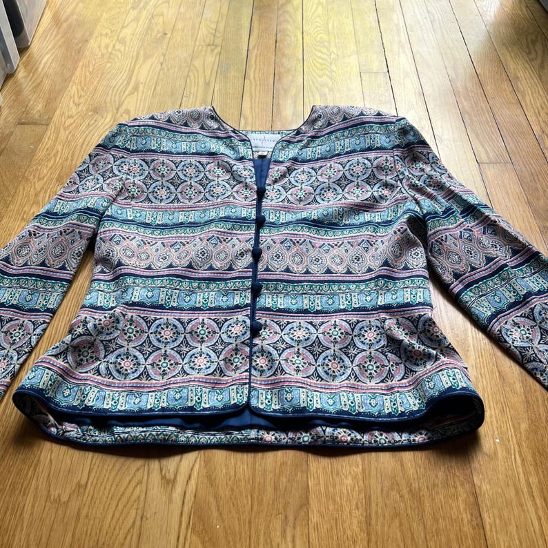 Peut inclure: Veste &agrave; motifs avec un col rond et des manches longues. La veste pr&eacute;sente une fermeture boutonn&eacute;e sur le devant et un motif color&eacute; avec des teintes bleues, roses et turquoise. Le tissu pr&eacute;sente un motif d&eacute;taill&eacute; et r&eacute;p&eacute;titif.