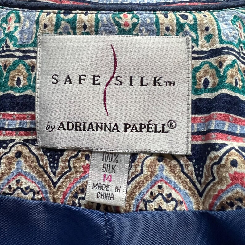 Peut inclure: Gros plan sur une &eacute;tiquette d'un v&ecirc;tement Safe Silk by Adrianna Papell. L'&eacute;tiquette est blanche avec du texte noir et une ligne incurv&eacute;e rouge. L'&eacute;tiquette indique que le v&ecirc;tement est 100% soie, taille 14, et fabriqu&eacute; en Chine. Le fond pr&eacute;sente un motif cachemire color&eacute;.