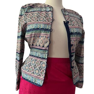 Peut inclure: Veste &agrave; motifs aux tons de bleu, rose et vert, pr&eacute;sent&eacute;e sur un mannequin. La veste a une encolure ronde et se ferme avec de petits boutons fonc&eacute;s. Elle est associ&eacute;e &agrave; une jupe rose vif. Le motif de la veste est complexe.