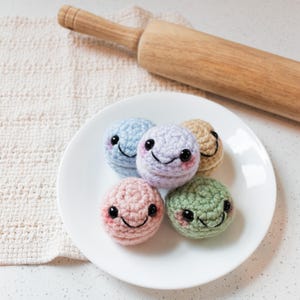 May include: A white plate holds five crocheted, smiling, round objects in pastel colors: light blue, lavender, tan, pink, and green. Each has black eyes and rosy cheeks. A wooden rolling pin rests in the background.