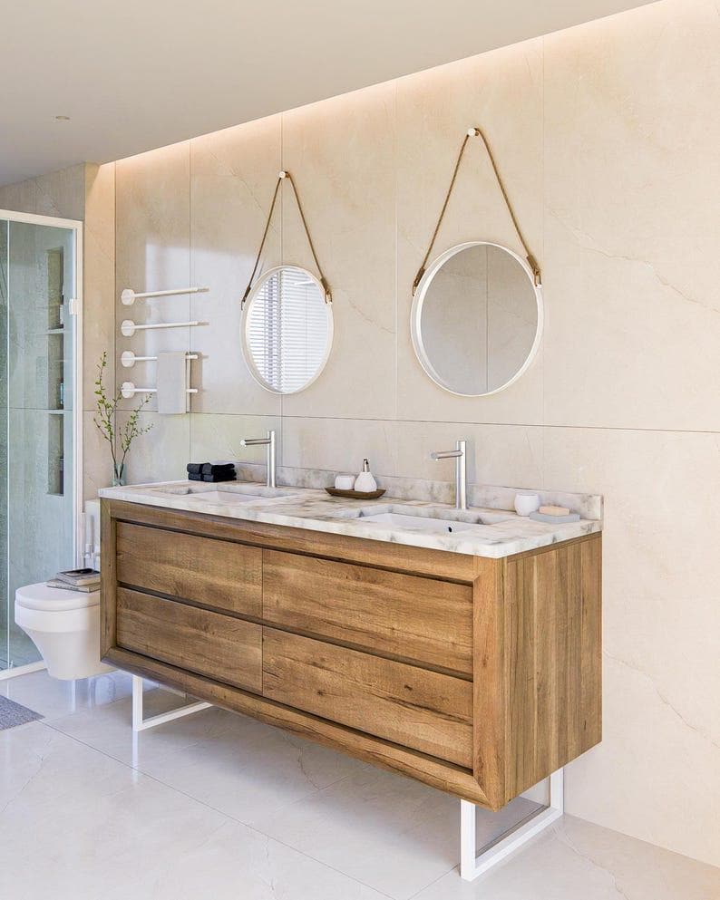 May include: A double vanity with a natural wood finish, four drawers, and a white marble countertop featuring two integrated sinks and silver faucets. Two round mirrors with white frames and brown leather hanging straps are mounted above the vanity.