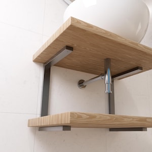 以下が含まれることがあります： 白いタイル張りの壁に取り付けられた、金属製のブラケットが付いた木製棚。棚は両側に2つの金属製のブラケットで支えられています。ブラケットは棚にネジで固定されています。