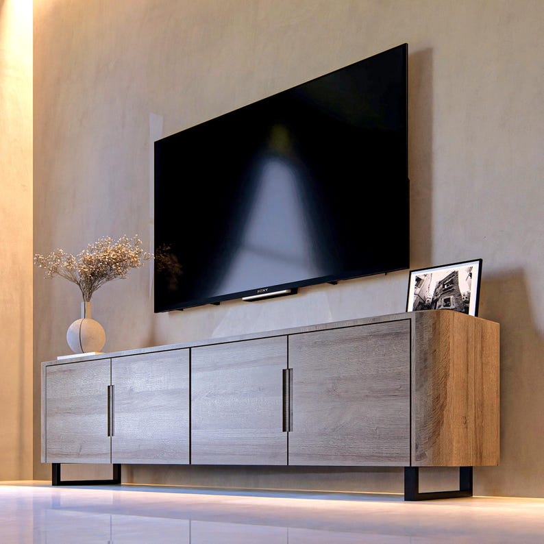 May include: A light brown wood grain media console with three cabinet doors and vertical metal handles, supported by black metal legs. A flat screen television is mounted on the wall above the console, next to a small white vase with dried flowers.