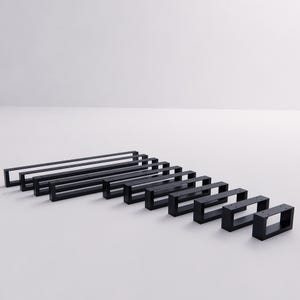 May include: A collection of ten rectangular black metal furniture legs arranged in a row from longest to shortest. Each leg features a hollow, open-frame design with pre-drilled holes along the top horizontal bar for mounting purposes.