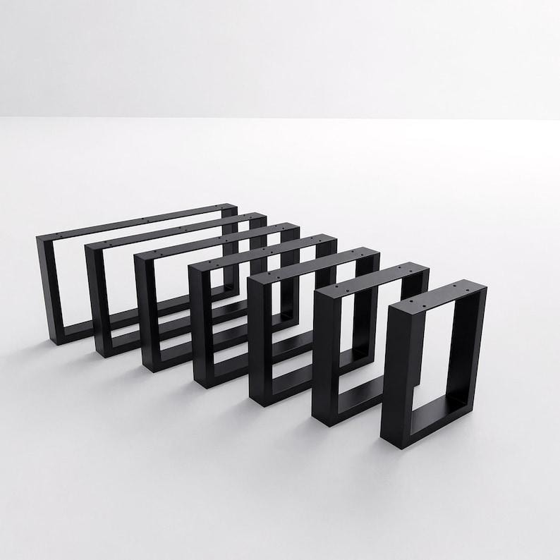 May include: A set of six rectangular black metal furniture legs arranged in a staggered row. Each leg features a hollow frame design with pre-drilled mounting holes along the top horizontal bar for attachment to a flat surface or furniture base.