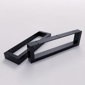May include: Two rectangular black metal furniture legs featuring a hollow center design. Each leg has a flat top mounting plate with pre-drilled circular holes for attachment, constructed from solid matte black steel with clean, sharp edges.