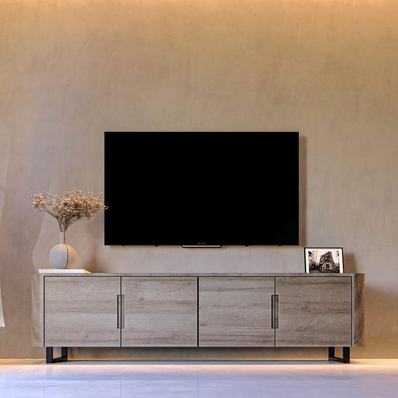 May include: A long, light-brown wooden media console with four cabinet doors and vertical metal handles, supported by black legs. A flat-screen television is mounted above the console, with a vase of dried flowers and a framed photo on top.
