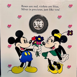 May include: A Valentine's Day card featuring Mickey Mouse and Minnie Mouse with a silver coin in a black case. The card has a pink and white background with hearts and the text "Roses are red, violets are blue, Silver is precious, just like you!"
