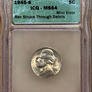 May include: A 1945-S nickel coin encased in a clear plastic holder. The coin features a profile of Thomas Jefferson and the words "IN GOD WE TRUST" and "LIBERTY 1945". A green label reads "ICG - MS64 Mint Error Rev Struck Through Debris".