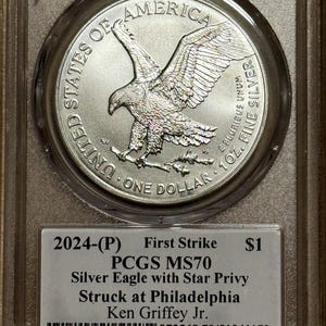 Peut inclure: Pi&egrave;ce d'argent am&eacute;ricaine de 2024, Silver Eagle, class&eacute;e PCGS MS70, avec un symbole d'&eacute;toile, dans un &eacute;tui de protection. La pi&egrave;ce repr&eacute;sente un aigle aux ailes d&eacute;ploy&eacute;es et le texte "UNITED STATES OF AMERICA". L'&eacute;tiquette indique "First Strike" et "1 oz. Fine Silver".