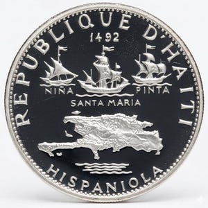 May include: A silver coin featuring three sailing ships labeled Niña, Santa Maria, and Pinta, with the text "REPUBLIQUE D'HAITI 1492". The coin also depicts a map of Hispaniola and the word "Hispaniola".