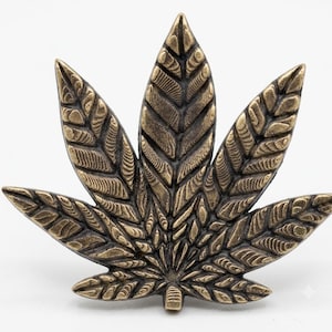 May include: A metallic bronze-toned pin shaped like a seven-pointed cannabis leaf. The surface features a detailed, raised texture with dark recessed lines between the segments, creating a dimensional appearance with a weathered, antique finish.