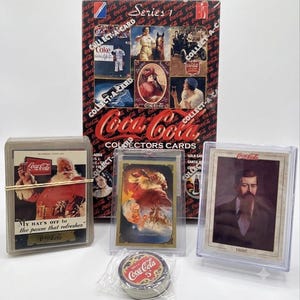 May include: A Coca-Cola collectors card box set featuring vintage imagery, accompanied by three individual trading cards in protective plastic cases and a small, round, metallic Coca-Cola branded token in a clear plastic wrapper.
