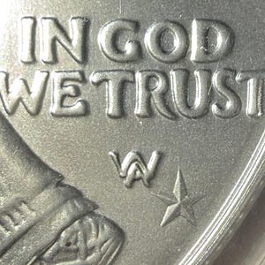 Peut inclure: Gros plan d'une pi&egrave;ce de monnaie argent&eacute;e avec les mots "IN GOD WE TRUST" en relief. La pi&egrave;ce pr&eacute;sente une marque d'atelier "W" et une &eacute;toile &agrave; cinq branches. Le bord de la pi&egrave;ce est visible.