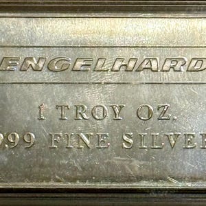 May include: A rectangular silver bullion bar encased in a clear protective plastic holder. The bar features the embossed text ENGELHARD, 1 TROY OZ., and .999 FINE SILVER. The metallic surface has a reflective finish with visible fine surface scratches.