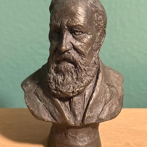 May include: A bronze-colored bust sculpture of a distinguished man with a full beard and a serious expression. The sculpture is a detailed representation of a man's head and shoulders, with a textured surface and a classic aesthetic.