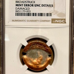May include: A copper-colored coin in a clear plastic NGC slab. The label reads (1983-2008) 1C MULTISTRUCK BROADSTRUCK MINT ERROR UNC DETAILS DAMAGED 8451179-003. The coin shows visible surface scratches and a distorted, multi-struck circular shape.