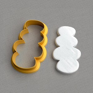 Puede incluir: Un cortador de galletas de plástico amarillo en forma de una serie apilada de formas redondeadas, junto a una galleta blanca texturizada con la misma forma. El cortador de galletas está sobre una superficie gris.