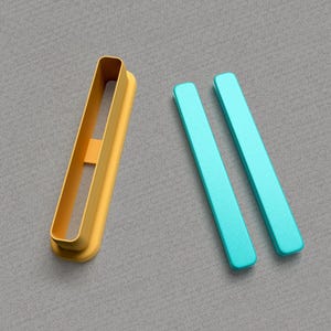Op de afbeelding: Twee turquoise plastic koekjesvormen in de vorm van parallelle lijnen, en één gele plastic koekjesvorm in de vorm van een rechthoek met een kleinere rechthoek uitgesneden in het midden.