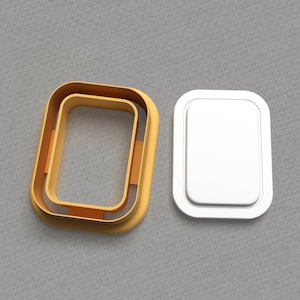 Peut inclure: Un emporte-pièce en forme de rectangle de couleur dorée avec un plus petit rectangle découpé au centre. L'emporte-pièce est ouvert et un couvercle en plastique blanc est à côté.