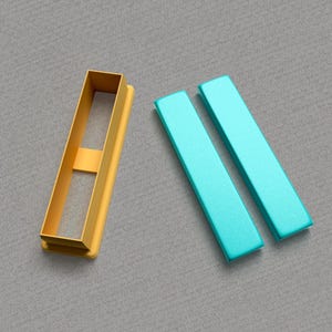 Peut inclure: Un ensemble de trois emporte-pièces, deux sont bleu turquoise et un est doré. L'emporte-pièce doré est en forme de rectangle avec un plus petit rectangle découpé au centre. Les deux emporte-pièces bleu turquoise sont en forme de longs rectangles.