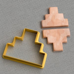 Può includere: Un tagliapasta in metallo dorato a forma di scala con cinque gradini. Il tagliapasta è su una superficie grigia testurizzata. Accanto al tagliapasta c'è un biscotto marrone chiaro che è stato tagliato con il tagliapasta.