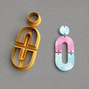 May include: Two cookie cutters, one shaped like the letter "i" and the other shaped like the letter "i" with a smaller circle in the middle. The "i" cookie cutter is made of blue and pink plastic, while the other cookie cutter is made of gold plastic.