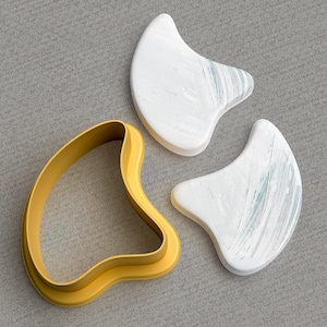 Puede incluir: Tres herramientas para el cuidado de la piel: dos piedras gua sha blancas y veteadas y un cortador de galletas amarillo y curvado. Las piedras gua sha tienen forma de lágrima curvada. El cortador de galletas tiene forma gruesa y redondeada.