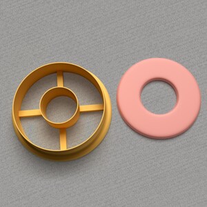 May include: A gold metal cookie cutter shaped like a donut with a smaller circle in the center. Next to it is a pink donut-shaped piece of fondant.