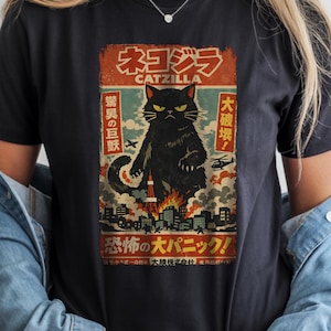 May include: Black t-shirt featuring a vintage-style graphic of a black cat labeled "CATZILLA" in Japanese text. The design includes a cat destroying a city with helicopters and explosions. The shirt is a classic crew neck.