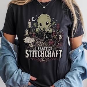 May include: A black t-shirt featuring a graphic illustration of a green octopus-like creature sewing with a needle and thread. The design includes candles, spools of thread, and scissors, with the text I PRACTICE STITCHCRAFT printed below.