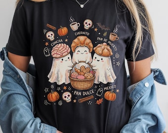 May include: A black t-shirt featuring a graphic of three white ghosts holding a woven basket filled with various breads. The design includes text labels such as Concha, Cuernito, Pan de Muerto, Pan Dulce, and Mexico, surrounded by pumpkins, skulls, and bats.