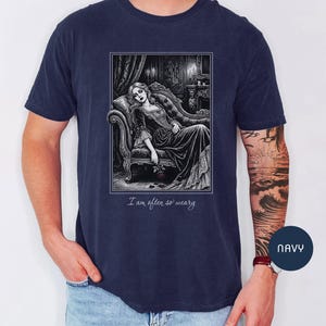 May include: Navy blue t-shirt featuring a black and white illustration of a woman reclining on a chaise lounge. The image includes the text "I am often so weary". The shirt is a size large.