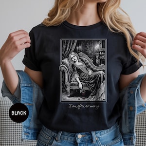 May include: Black t-shirt featuring a black and white illustration of a woman reclining on a chaise lounge in a gothic setting. The text "I am often so weary" is printed below the image. The shirt is a dark color.