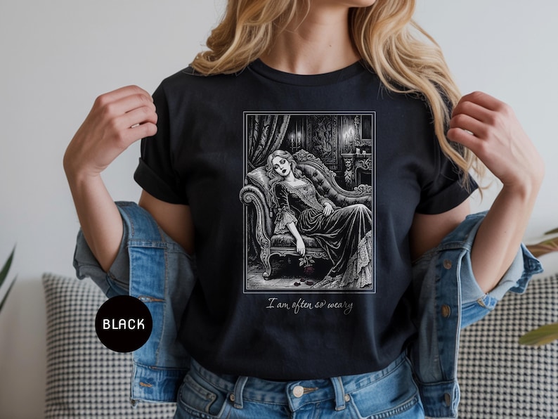 May include: Black t-shirt featuring a black and white illustration of a woman reclining on a chaise lounge in a gothic setting. The text "I am often so weary" is printed below the image. The shirt is a dark color.