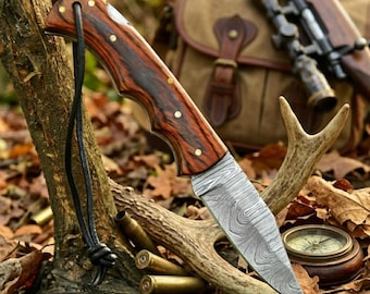 Puede incluir: Una navaja plegable de acero de Damasco con mango de madera oscura y remaches de latón. El cuchillo tiene un cordón de cuero y se exhibe con equipo de caza, incluyendo un rifle, una bolsa y una brújula. El cuchillo es de WBSmith Knives.