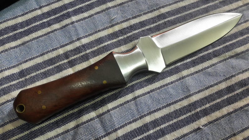 May include: A brown handled knife with a silver blade. The blade is sharp and pointed. The handle is made of wood and has a smooth finish. The knife is lying on a striped fabric surface.