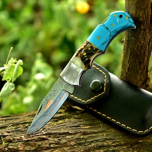 May include: A folding knife with a turquoise handle and a patterned blade rests on a wooden surface. The knife has a leather sheath with yellow stitching. The blade has a unique pattern and the handle has a decorative design.