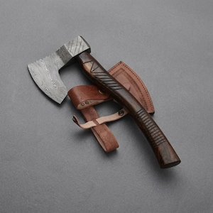 May include: A hand axe featuring a patterned Damascus steel blade and a dark brown wooden handle with carved geometric grip details. The axe is accompanied by a tan leather sheath with metal rivets and a strap, designed to cover the blade edge.
