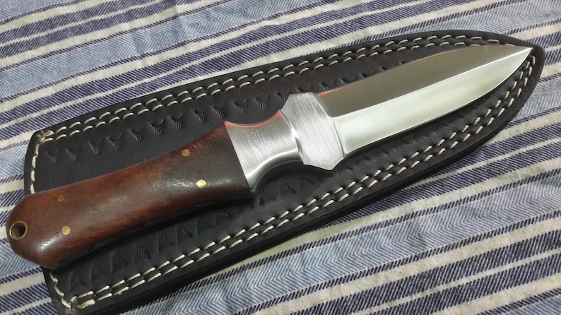 May include: A brown handled knife with a silver blade and a black leather sheath. The sheath has white stitching around the edges.