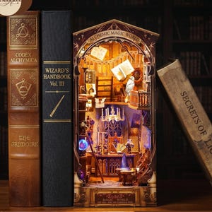 May include: A book nook diorama titled Alchemic Magic World, flanked by three faux books titled Codex Alchymica, Wizard's Handbook Vol. III, and Secrets of Light. The interior features a miniature library with glowing lights, floating books, and a white owl.