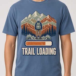 May include: A blue short-sleeved t-shirt featuring a graphic of a hiker with a backpack standing before mountains and pine trees. Below the illustration is a partially filled orange loading bar and the text "TRAIL LOADING" in bold letters.