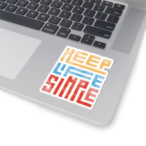 May include: A white sticker with the words "KEEP LIFE SIMPLE" in a stacked design. The text is in orange, blue, and red, with a distressed, textured appearance. The sticker is on a laptop.