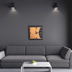 Könnte beinhalten: Eine quadratische Wanduhr aus Holz mit hellbraunem Zifferblatt und schwarzen Zeigern. Die Uhr hat einen schwarzen Rahmen und das Wort "three" ist auf der rechten Seite aufgedruckt. Die Uhr ist an einer grauen Wand über einem grauen Sofa befestigt.