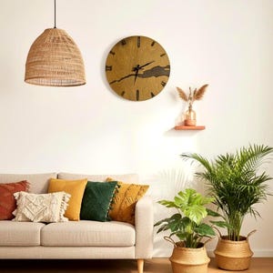 May include: A living room scene featuring a beige sofa with decorative pillows in shades of orange, yellow, and green. A large wooden clock hangs on the wall, and a woven pendant light hangs from the ceiling. Potted plants in woven baskets add a natural touch.