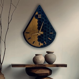 Könnte beinhalten: Eine tropfenförmige Wanduhr aus Holz mit zweifarbigem Design. Die linke Seite ist aus Naturholz, während die rechte Seite marineblau mit einem Wabenmuster ist. Die Zeiger und die römische Ziffer XI sind ebenfalls aus Holz.