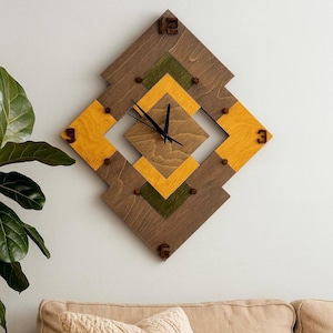 May include: Diamond-shaped wooden wall clock with a geometric design. The clock features brown, yellow, and green wood panels. Black clock hands and wooden numerals at 3, 6, 9, and 12 complete the design. The clock is mounted on a white wall.
